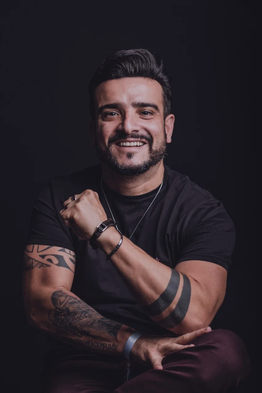 Retrato de Rômulo Barale, sorrindo, braços cruzados, sobre fundo escuro.
